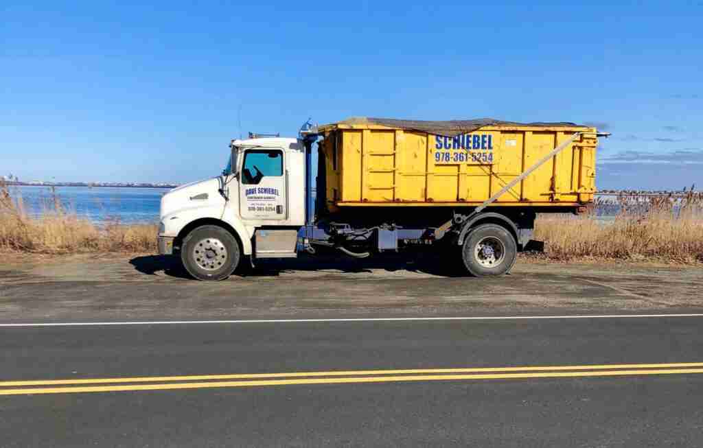 A white schiebel waste truck carrying a large yellow roll off dumpster